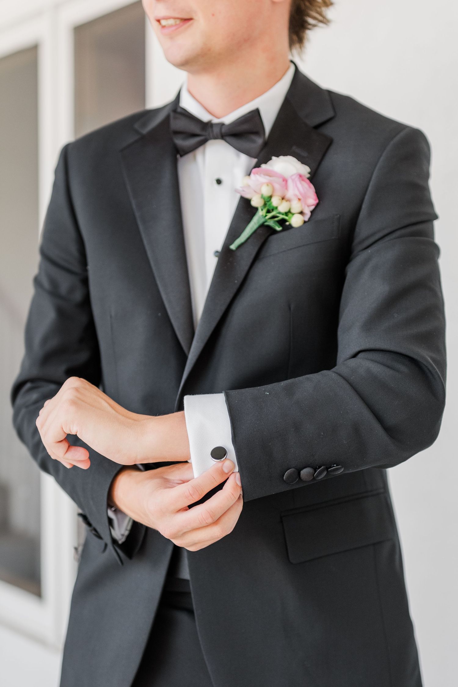 Groom adjusts cufflinks and boutonniere while getting ready in classic black tuxedo.