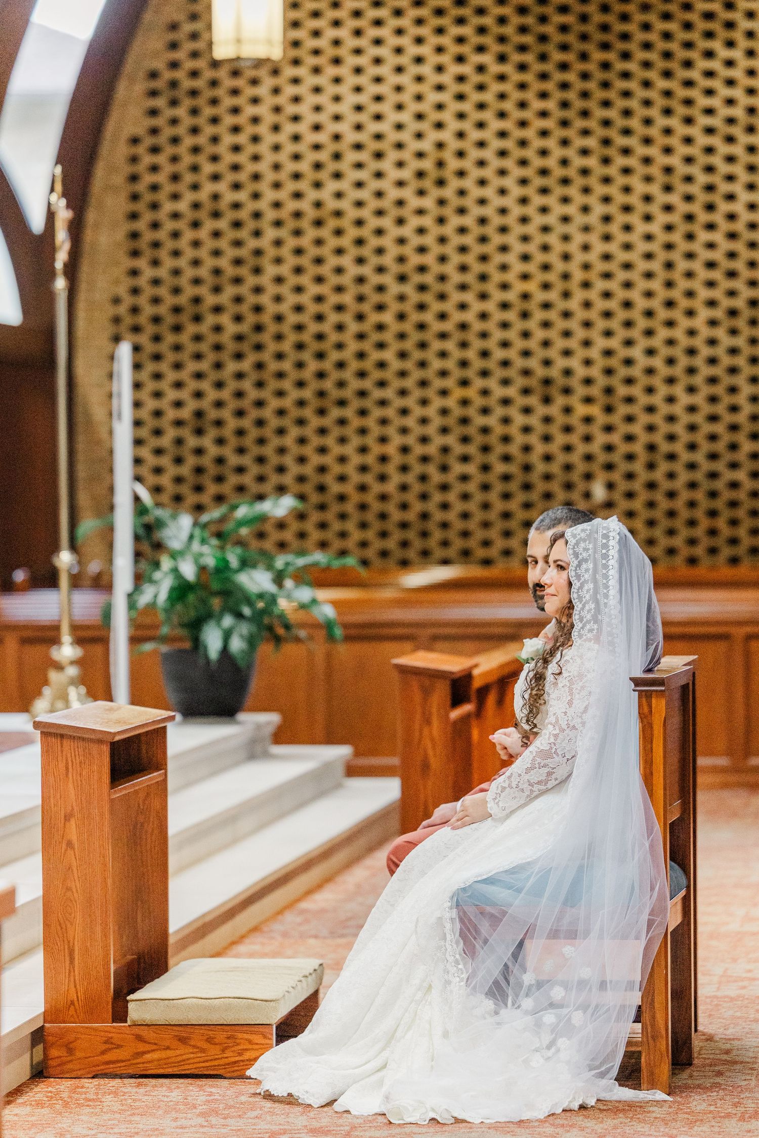 A bride in a white lace wedding dress sits on wooden church pews while her veil flows elegantly behind her.