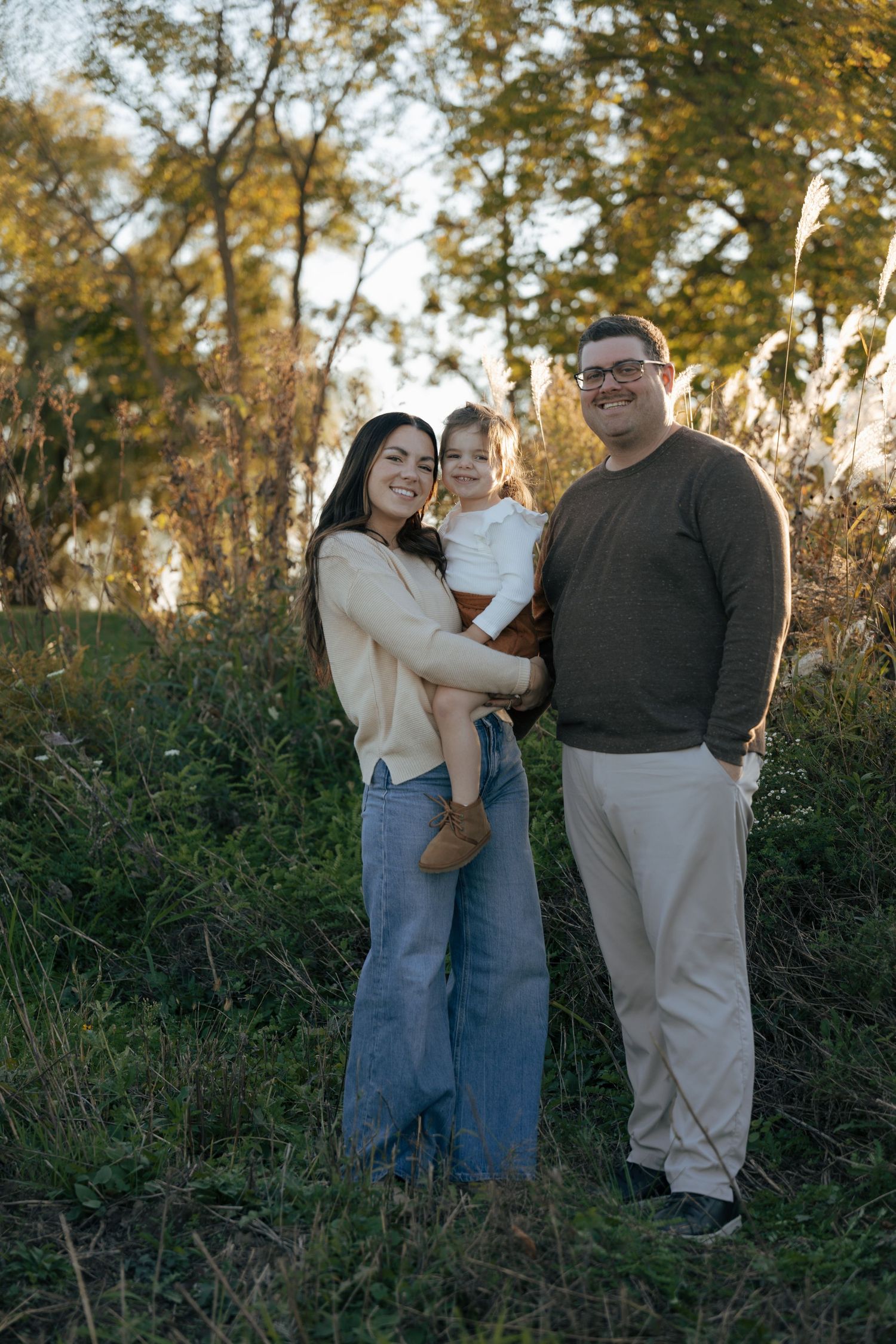 Family standing together in a field during golden hour autumn sunset with tall grass and trees in background.