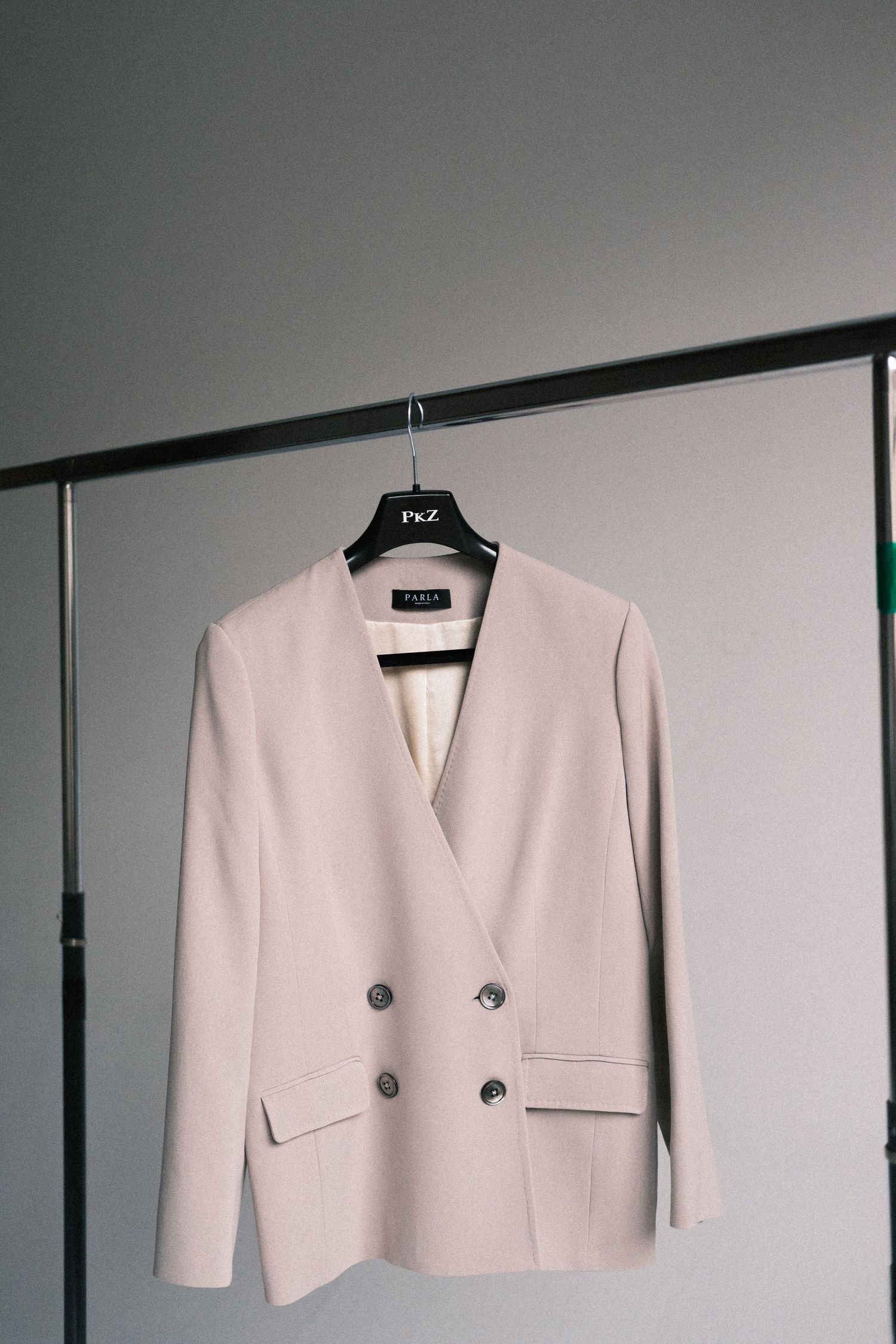 A white cropped blazer jacket with silver buttons displayed on a black hanger against a gray wall.
