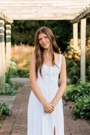 A woman in a white sundress stands in a garden path lined with brick pavers and lush greenery.