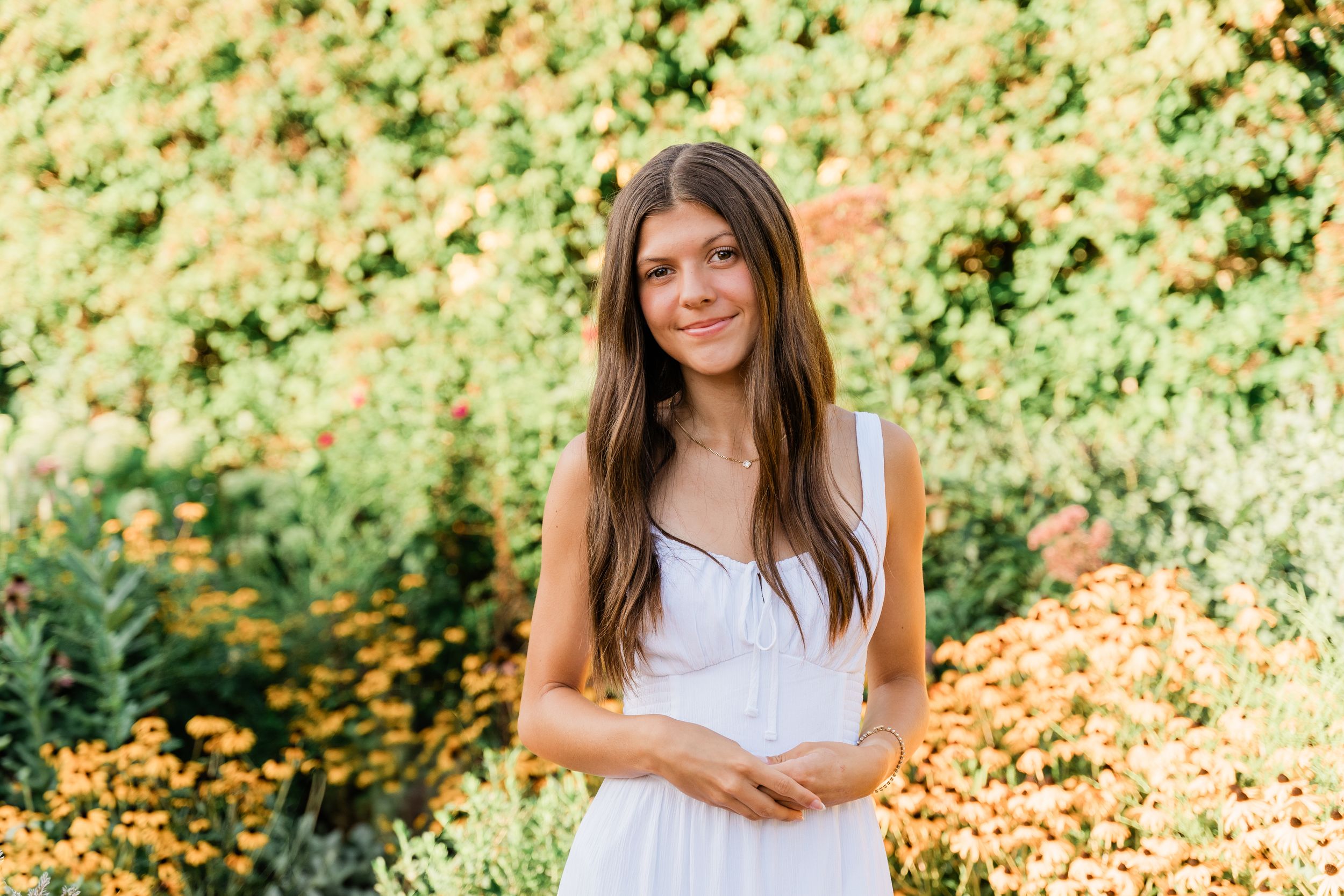 A person in a white dress stands in a sunny garden with golden bokeh lighting and green foliage in the background.