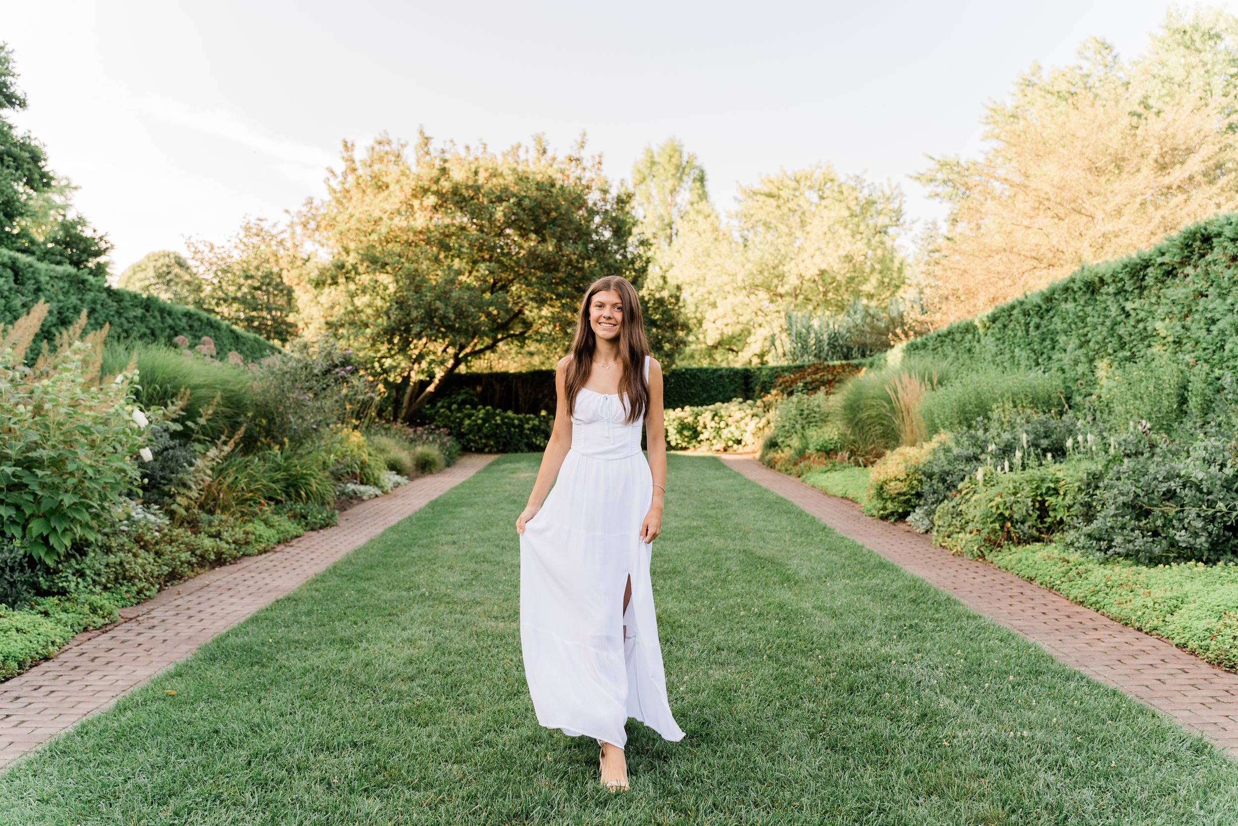A white sundress flows gracefully in a sunlit garden path surrounded by lush greenery.