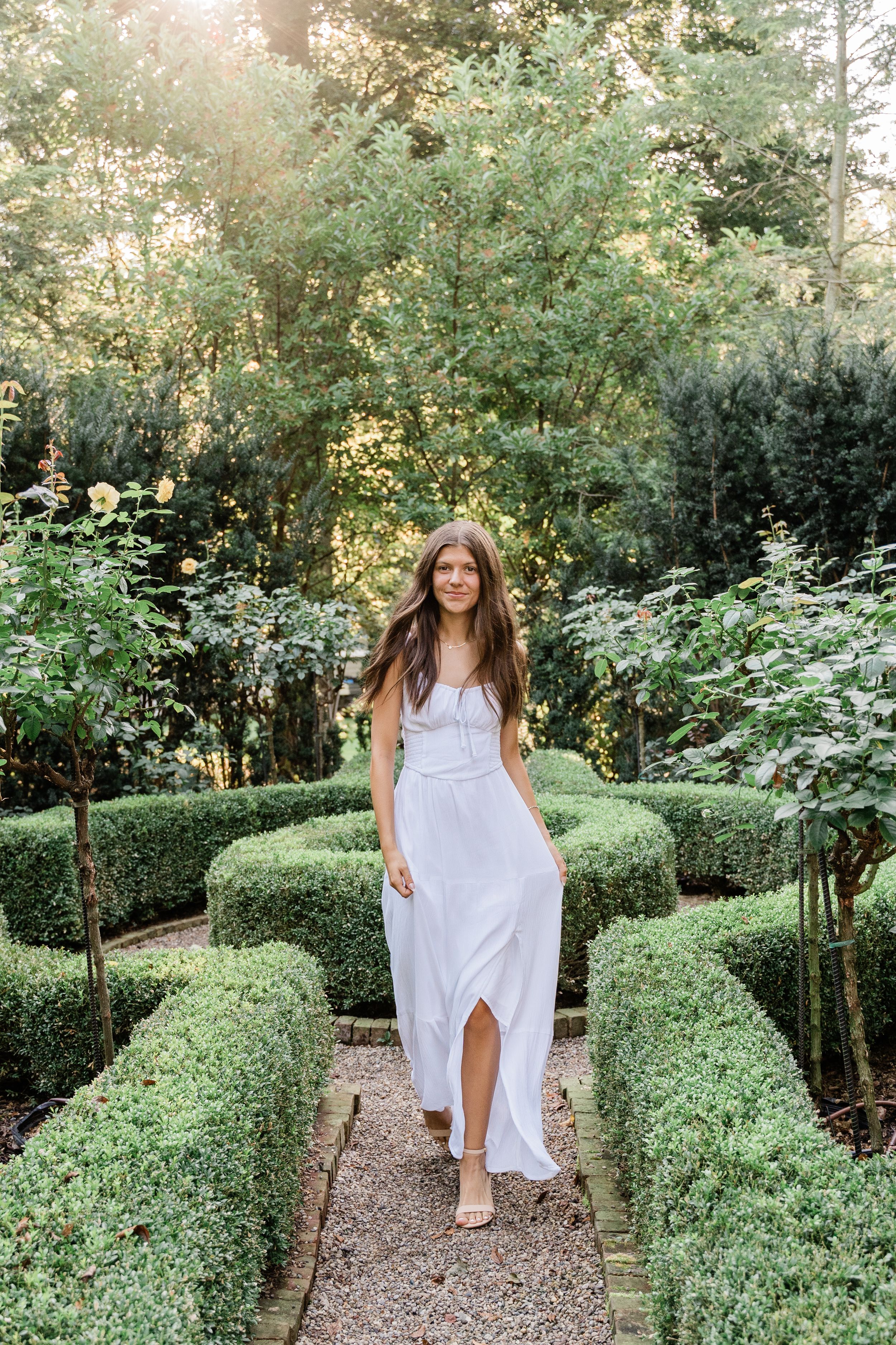 A person in a flowing white dress walks along a manicured garden path lined with neatly trimmed boxwood hedges.