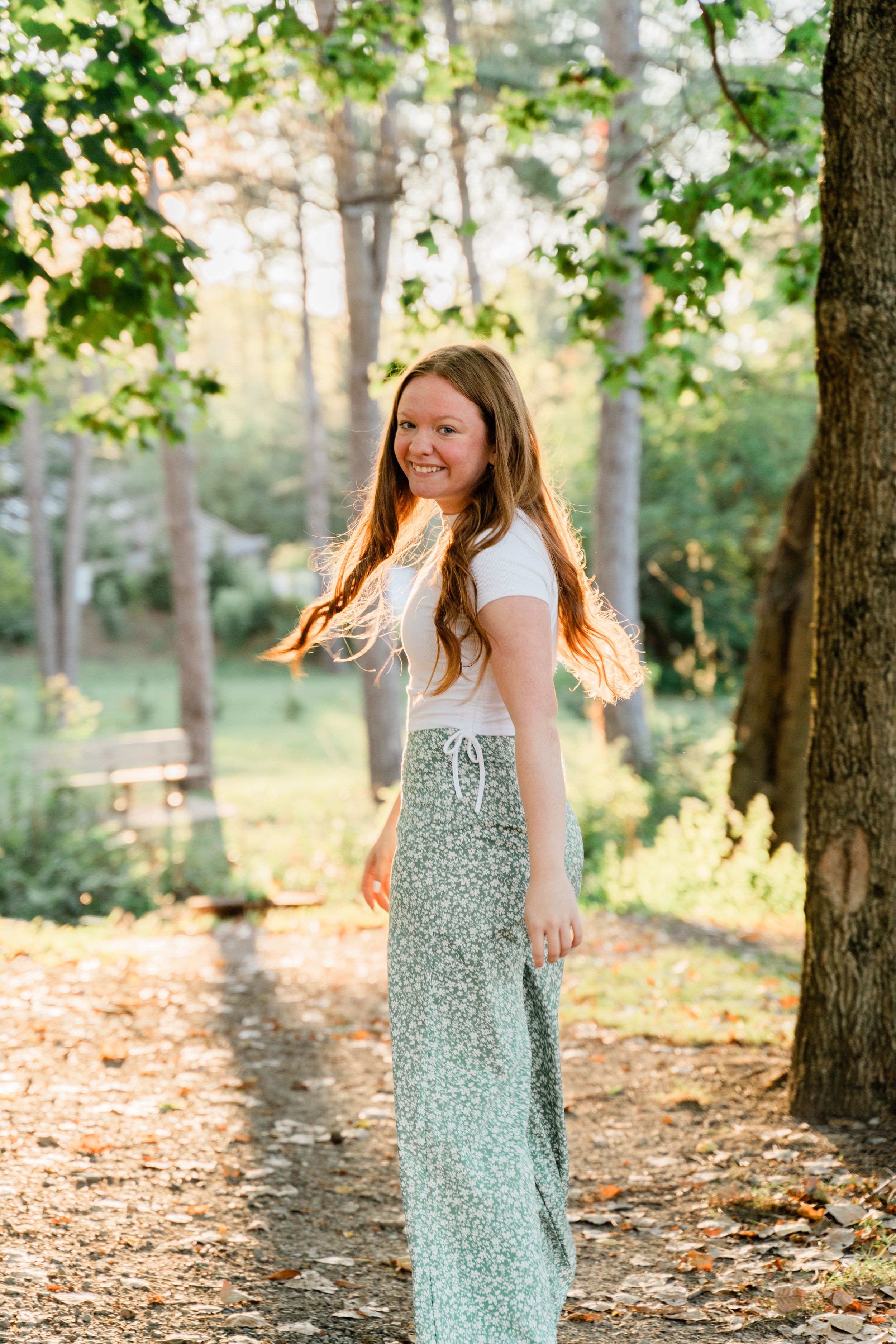 A woman in a floral dress and white top twirls playfully among trees in a sunlit forest setting.