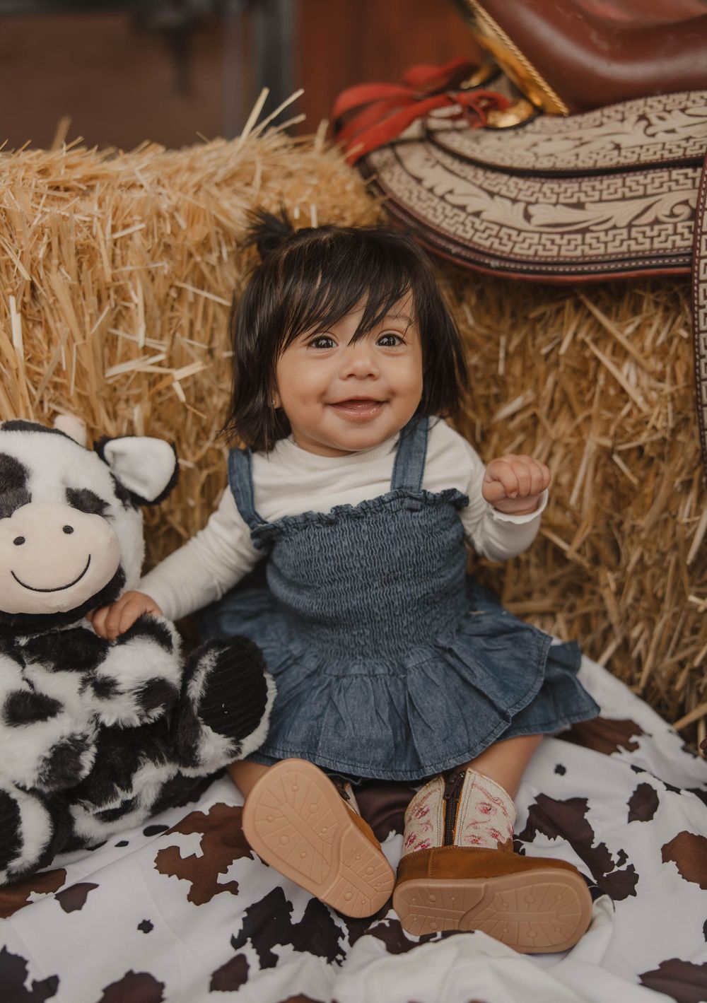 Child sitting on hay bales with stuffed cow toys and western decor.