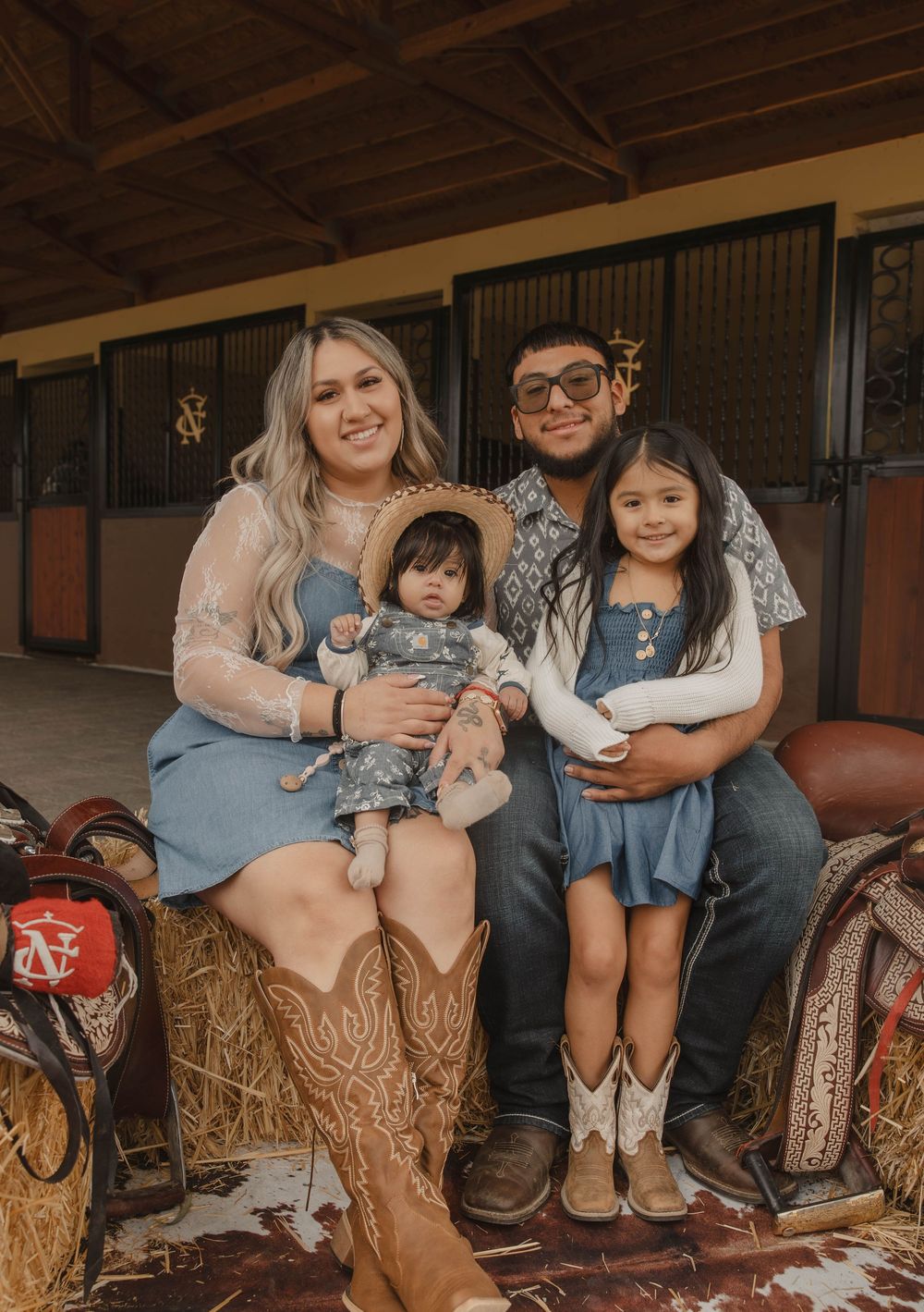 Family wearing western boots sits together on hay bales in a rustic barn setting.