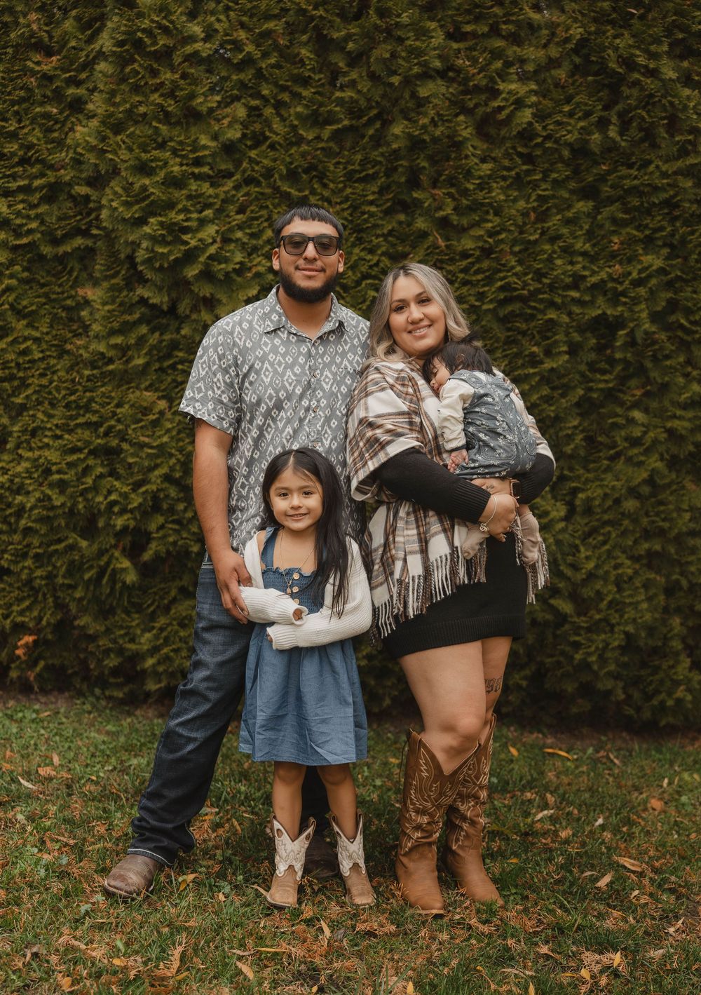 A family poses together in front of a green hedge wearing casual fall attire.
