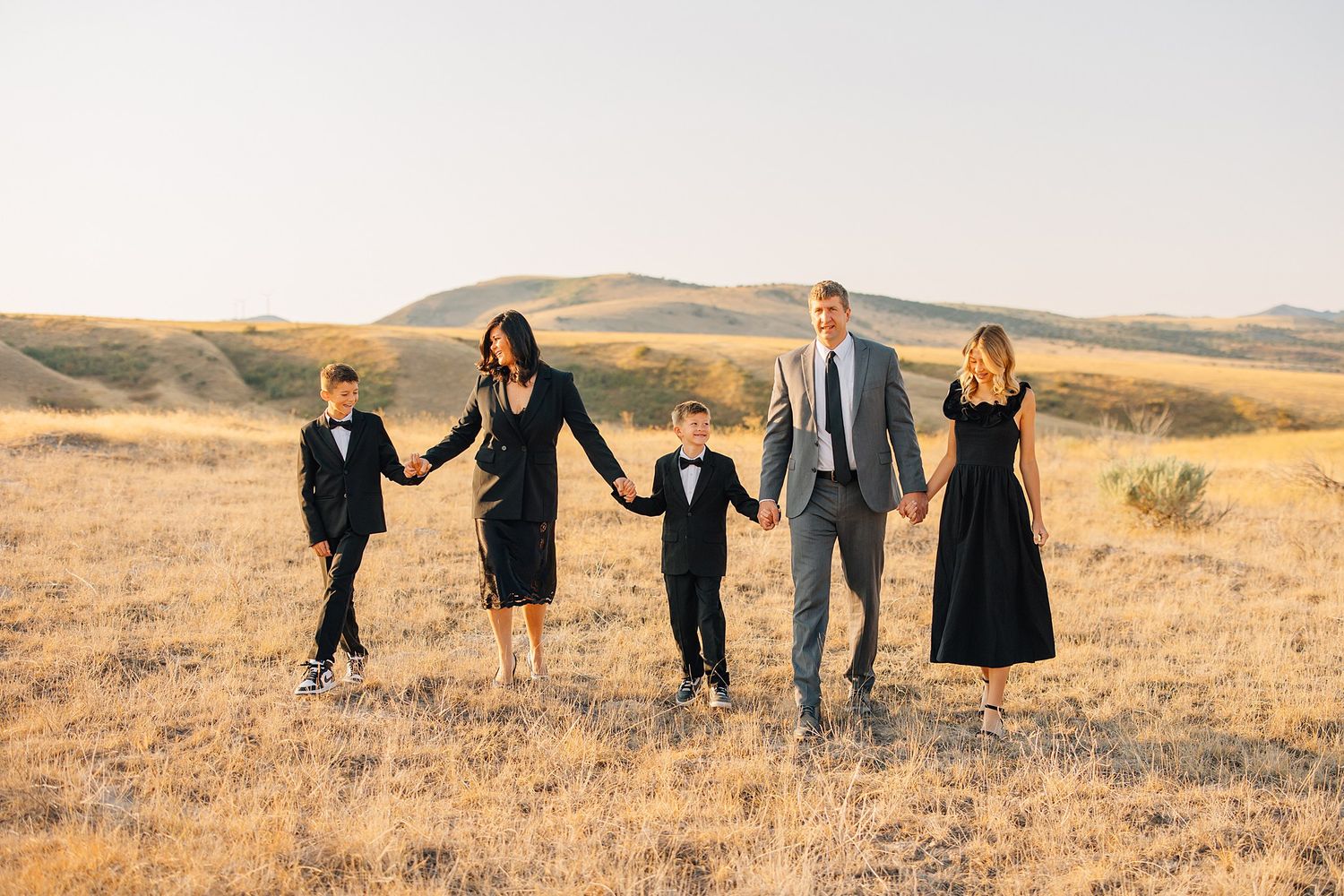 A family of five holds hands while walking through a golden autumn field at sunset with rolling hills in the background.