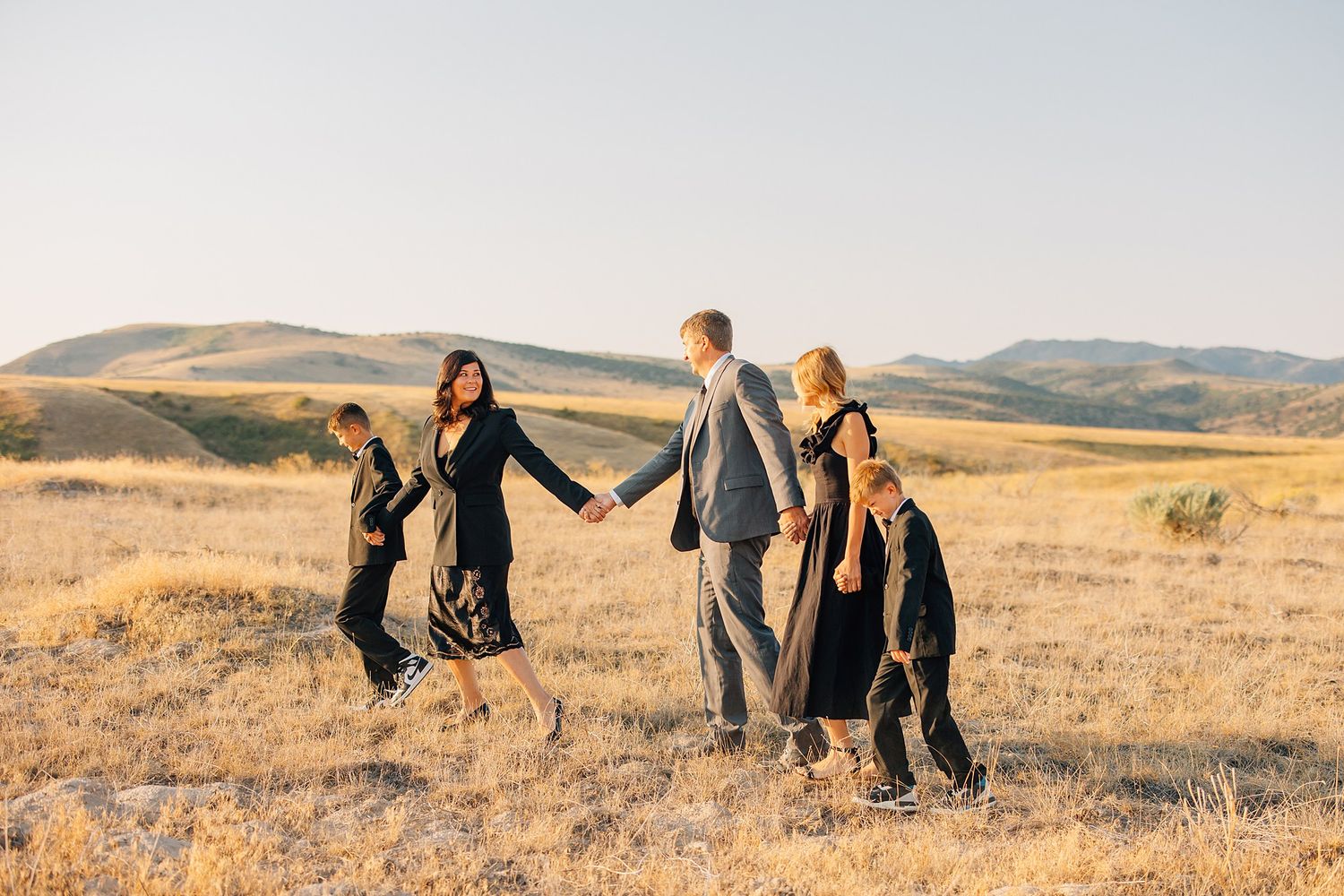 People holding hands while walking together through a golden field with rolling hills in the background at sunset.