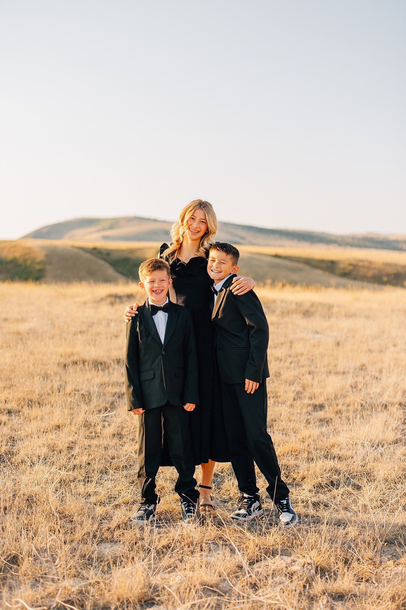 Three children in formal black attire laugh and play together in a golden grassy field at sunset.
