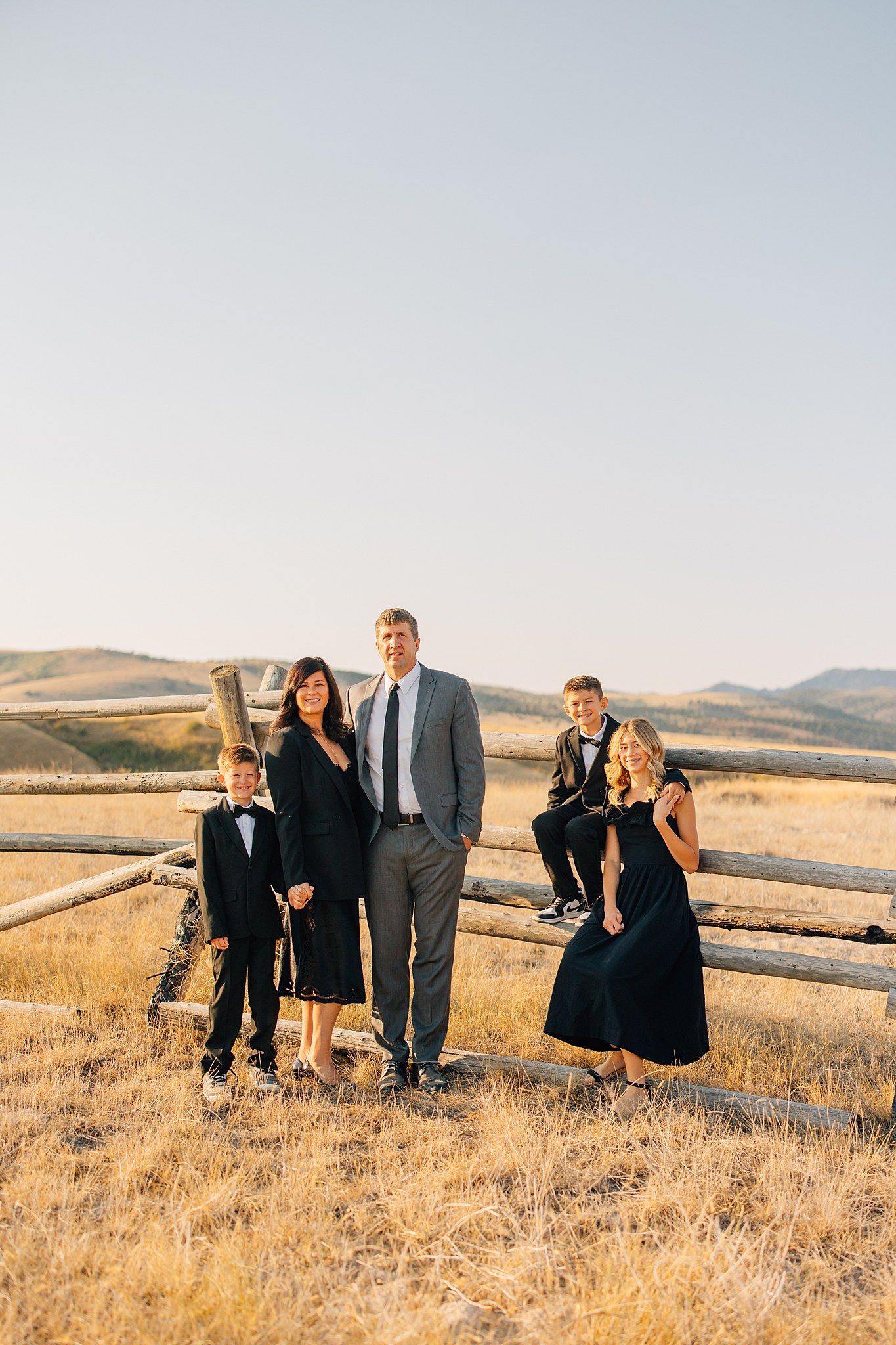 Family in formal attire poses together on a hillside with wooden fence and golden grass at sunset.