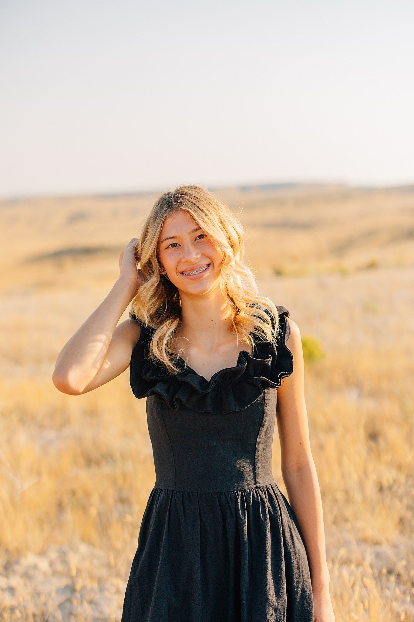 Woman in black dress poses in golden field at sunset.