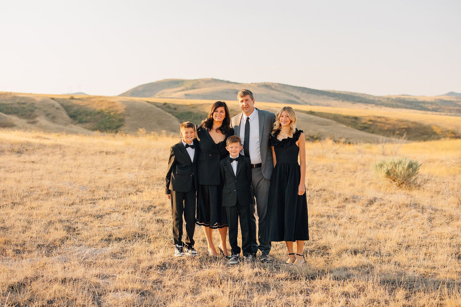 A family poses together in formal attire on a golden hillside at sunset during a photo session.