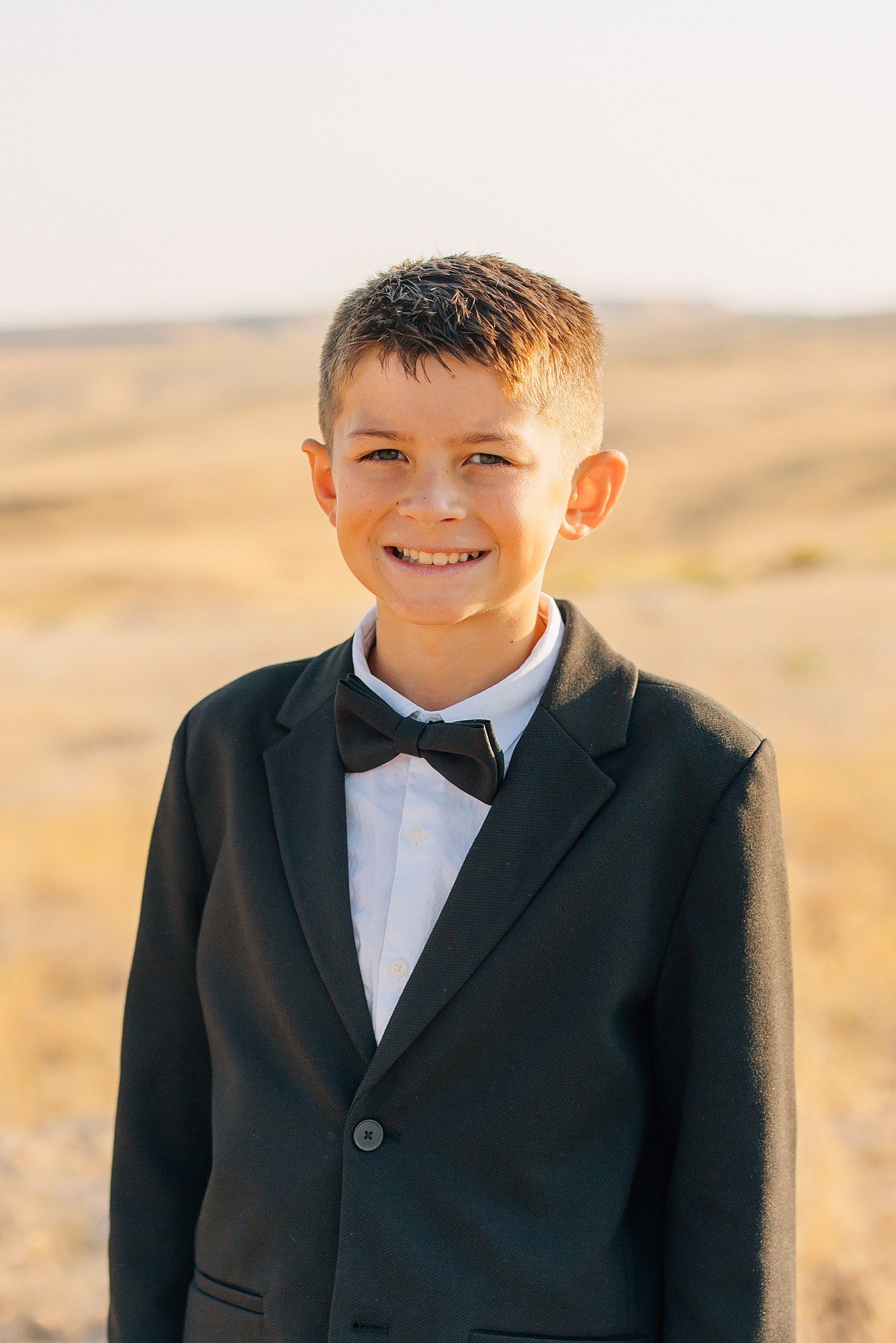 Young boy in formal black tuxedo and bowtie smiling against desert landscape at sunset.