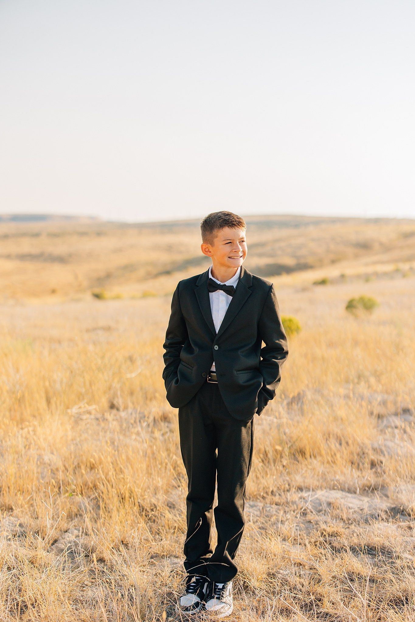 Child in black suit poses playfully in golden field with rolling hills in background.