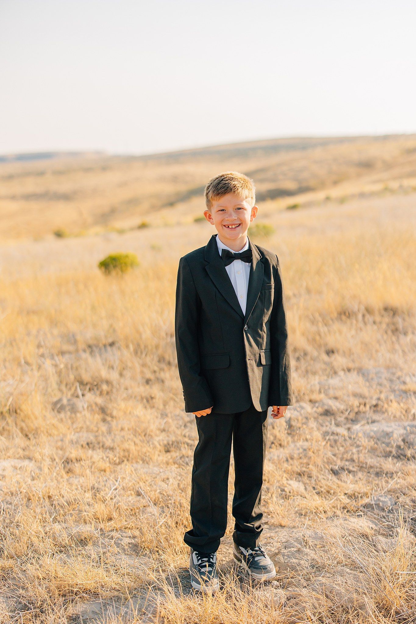 A small child wearing a formal black suit stands in a golden field during sunset.