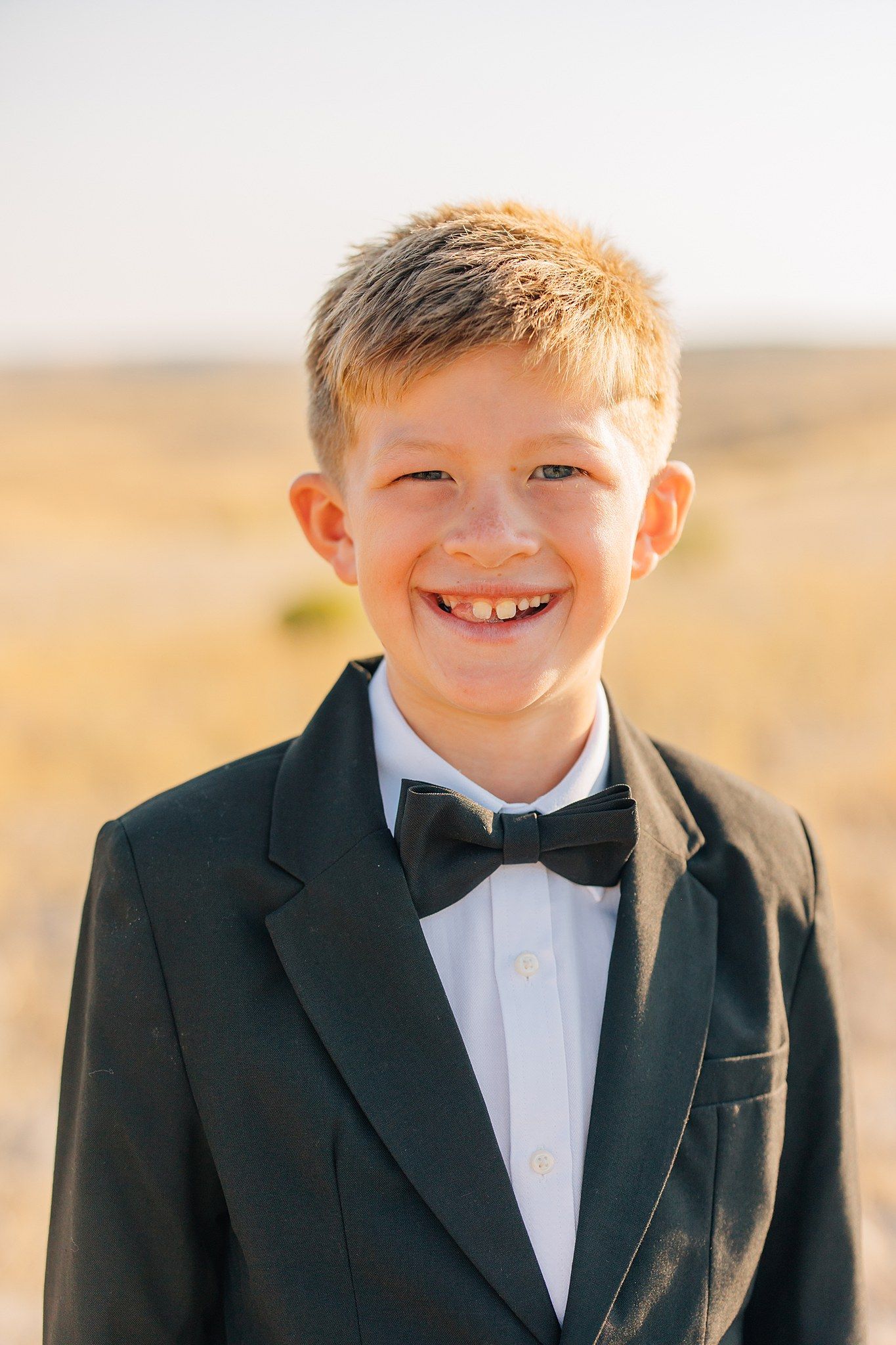 Young child in formal black tuxedo and bowtie smiling outdoors against a golden sunset background.