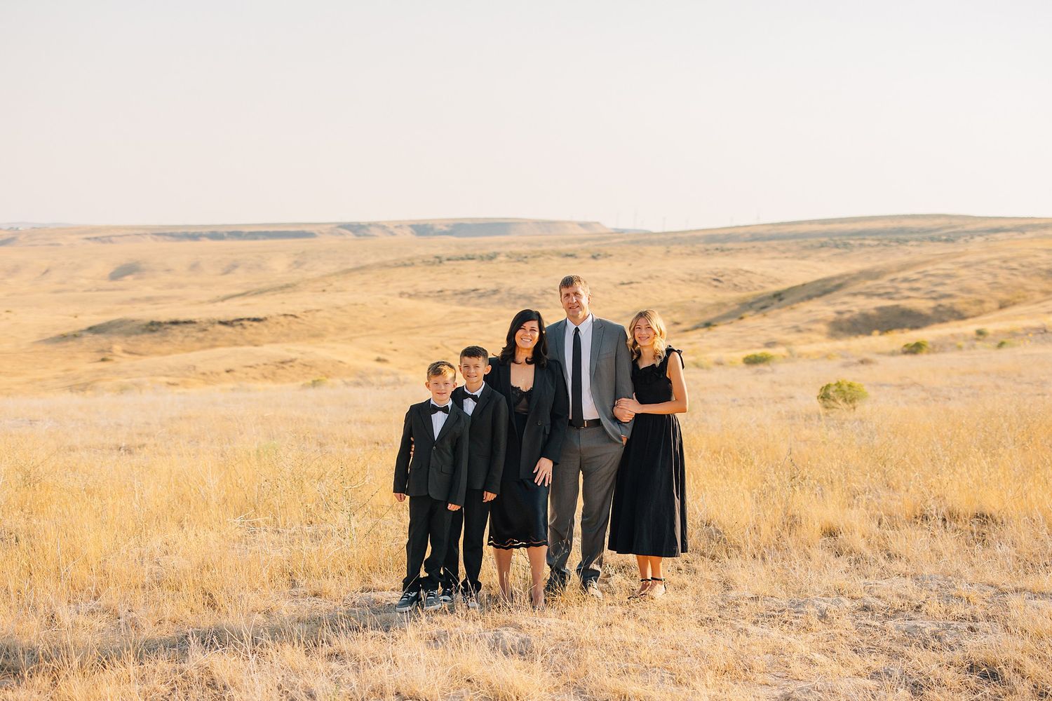 A panoramic series of family portraits in a golden prairie landscape at sunset with rolling hills in the background.