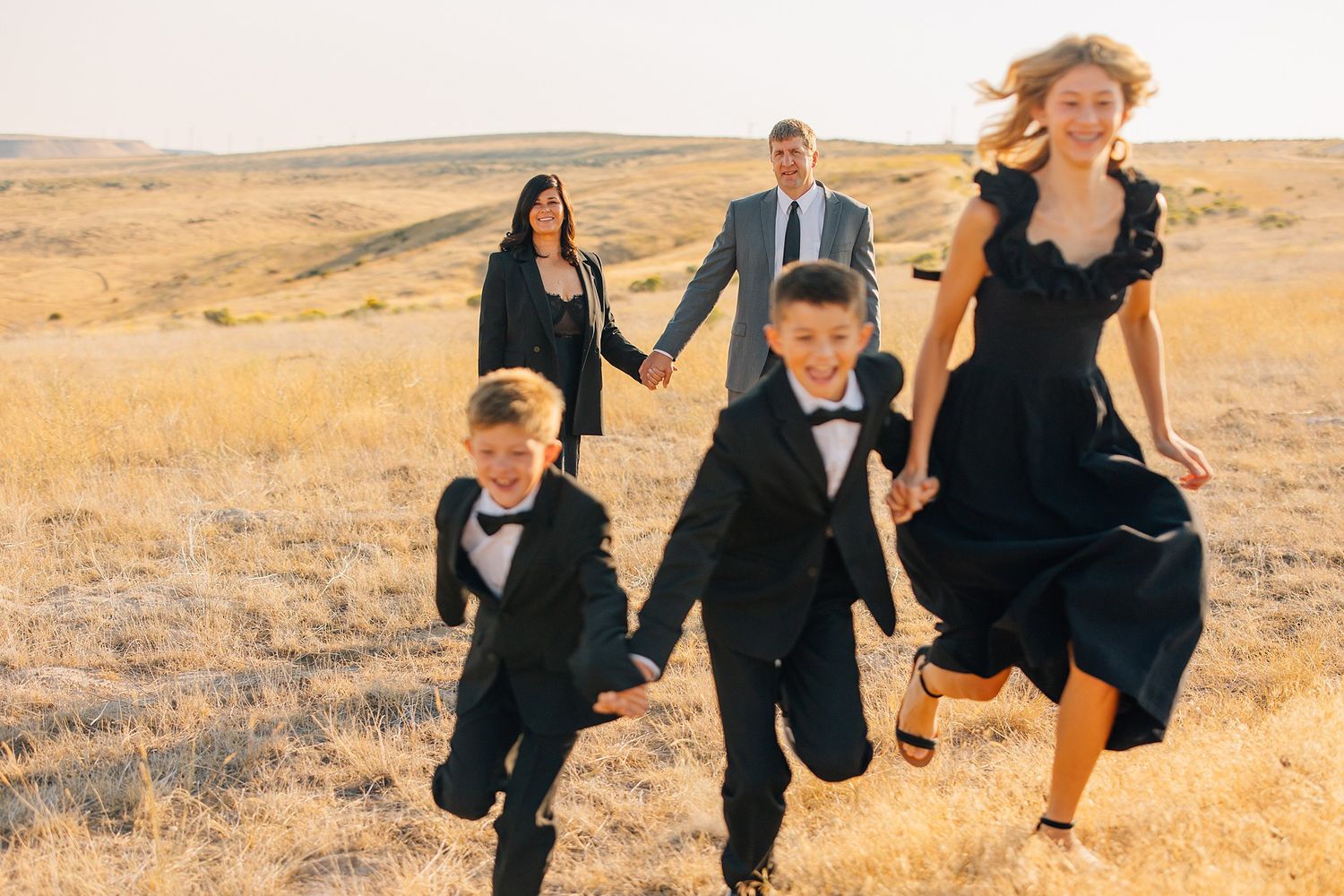 A group in formal black attire runs joyfully through a golden wheat field at sunset.