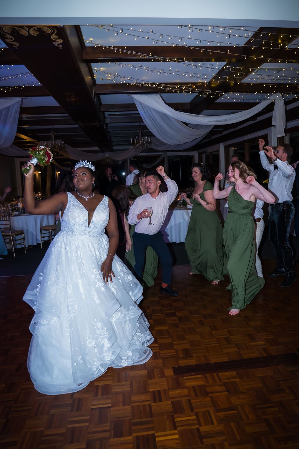 Wedding reception guests dancing and celebrating on a wooden dance floor under twinkling lights.