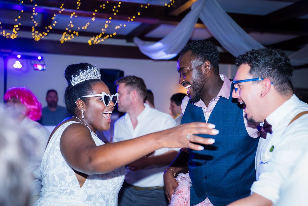 Wedding reception party scene with people dancing and celebrating under decorative string lights and draped fabric.