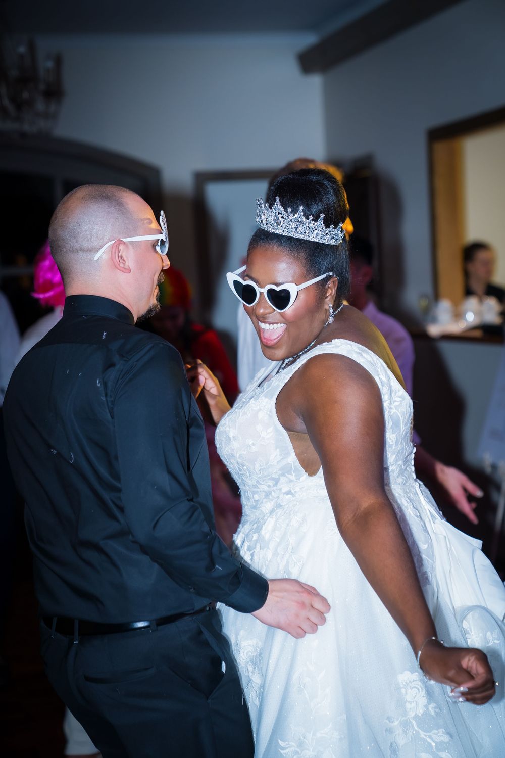 A couple in formal attire dance together at a wedding reception, with the bride wearing a white dress and sunglasses.