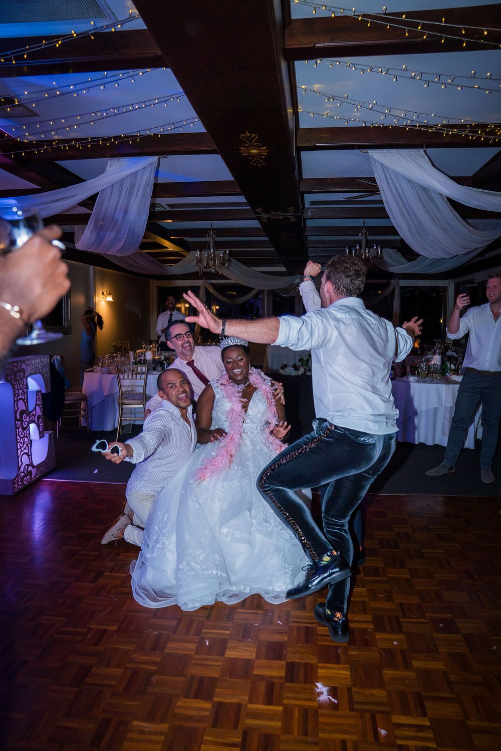 Wedding guests perform synchronized dance moves on a wooden dance floor under twinkling string lights at an indoor reception.