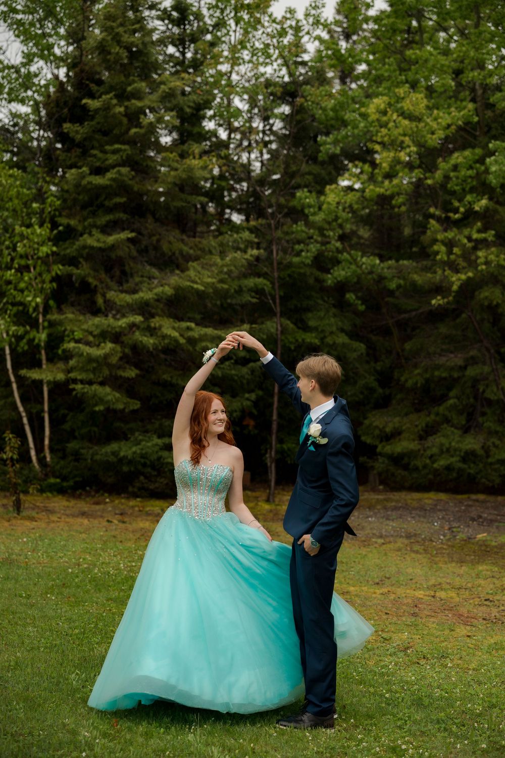 A couple dances outdoors, woman in teal ballgown twirling, man in navy suit, surrounded by lush green trees.