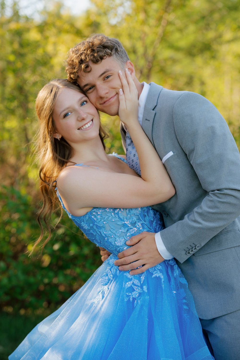 A smiling teenage couple in prom attire, girl in blue gown and boy in gray suit, posing outdoors.