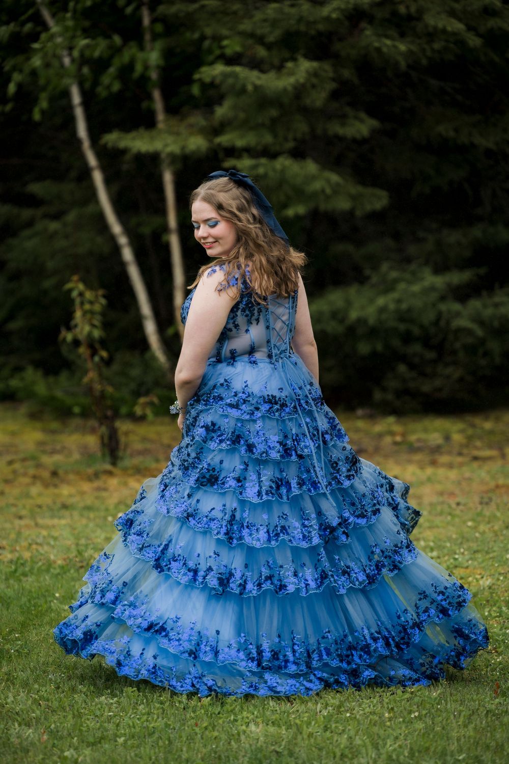 Woman twirling in a tiered blue floral lace ball gown outdoors in a lush green garden setting.