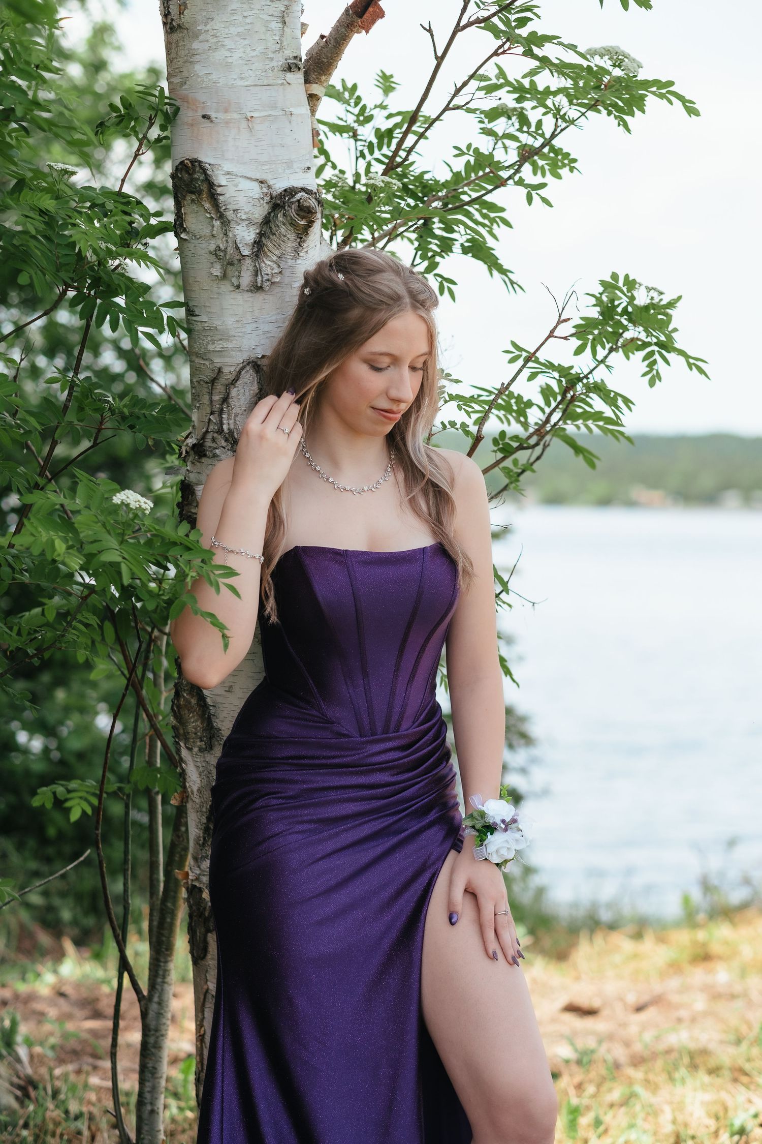 A woman in a purple dress poses elegantly against a birch tree by a lake on a sunny summer day.