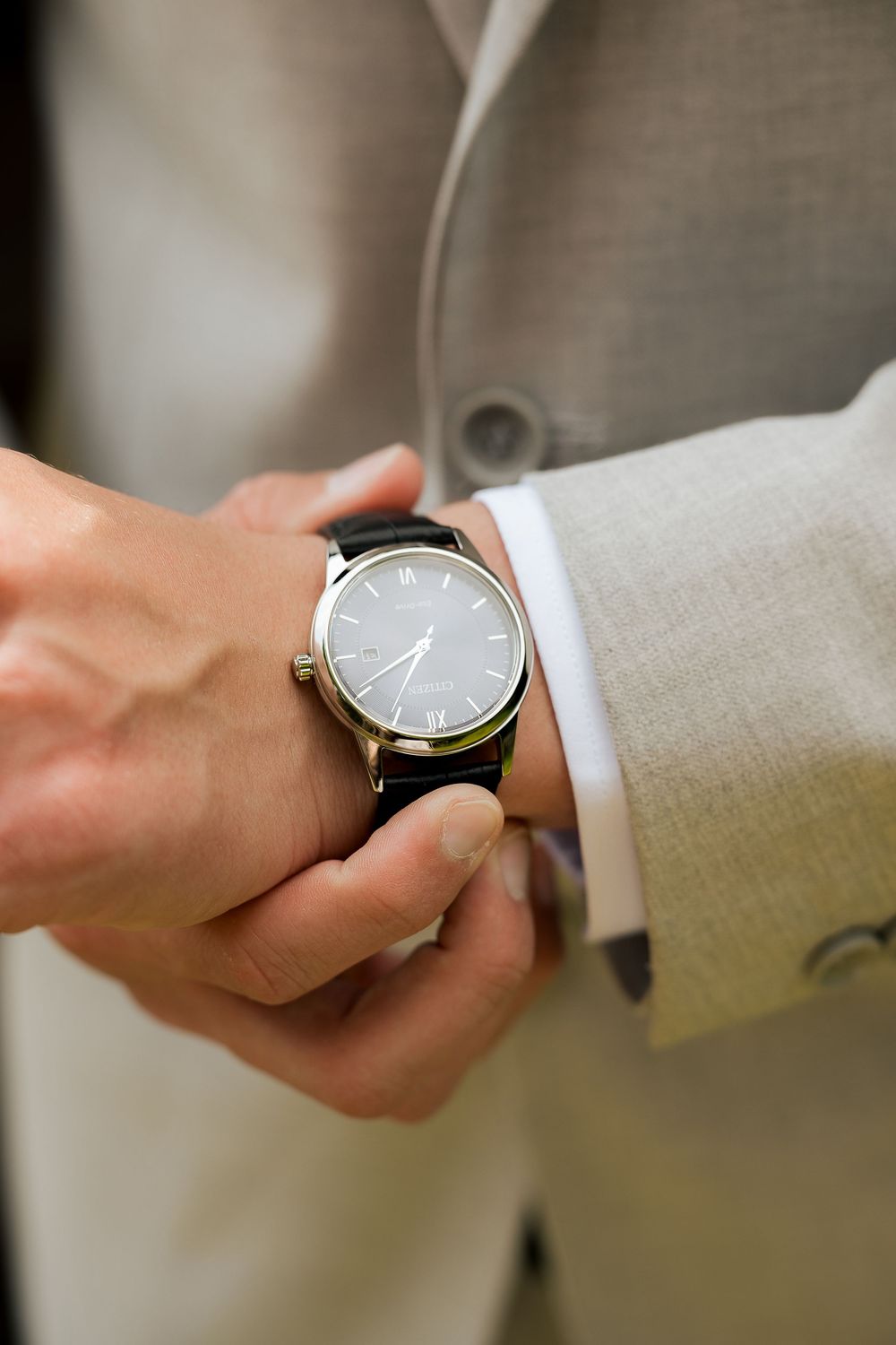 Man in beige suit adjusting a classic black dial wristwatch with leather strap on his wrist.