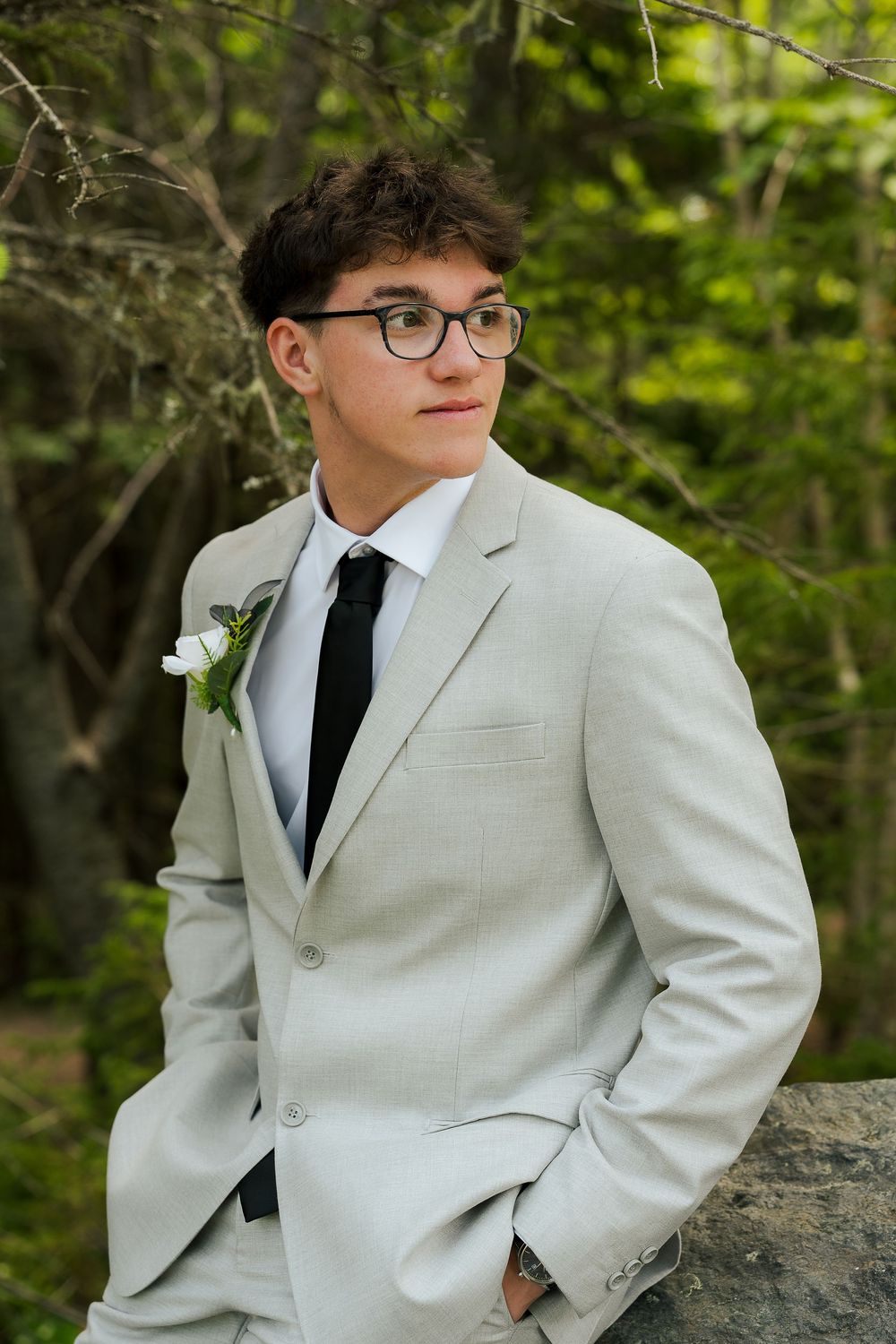 Young man in light gray suit with black tie and white boutonniere, wearing glasses, posing outdoors near green trees.