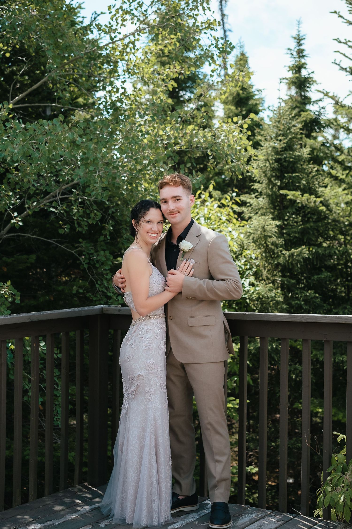 A couple shares a romantic dance moment on an outdoor wooden deck surrounded by evergreen trees in the sunlight.