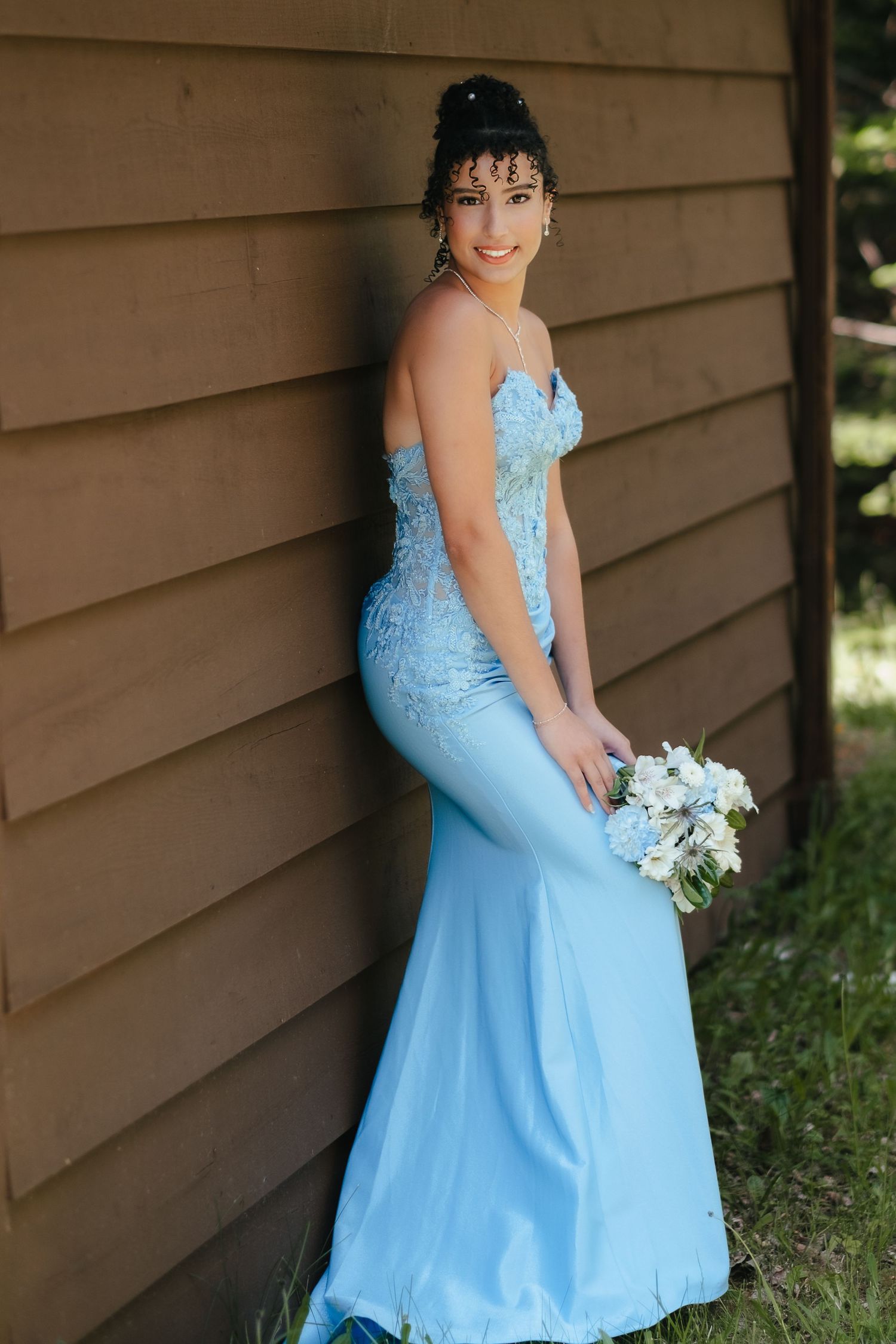 A person in a light blue formal dress poses against a brown wall while holding a white floral bouquet.