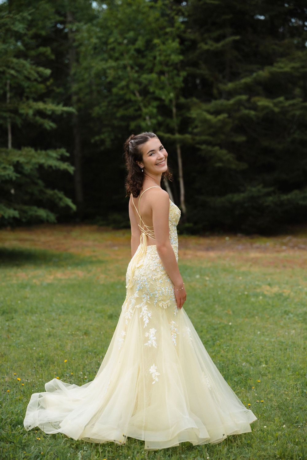 Young woman in elegant pale yellow floral gown smiling outdoors in a lush green forest setting.
