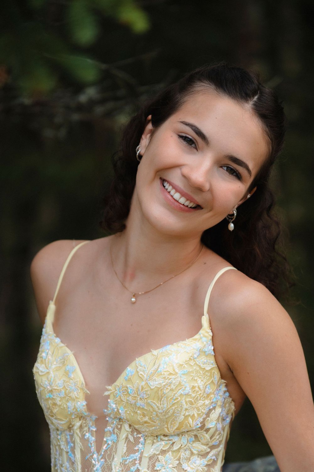 Smiling young woman in a yellow floral embroidered dress with pearl earrings and necklace outdoors.