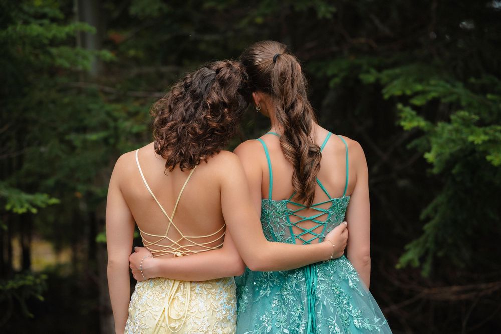 Two young women in elegant backless prom dresses embracing outdoors with forest background.
