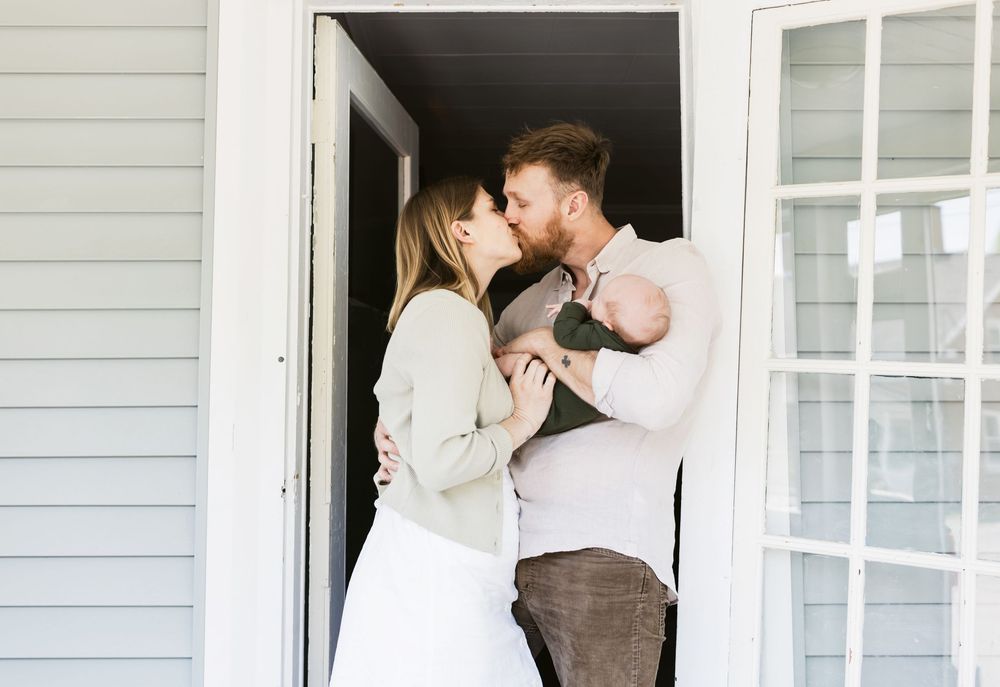 A couple kisses in the doorway of their porch for a lifestyle newborn shoot in Berwick, Maine. Berkshires newborn photographer.
