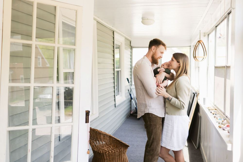 A happy couple wearing pastel and neutral colors gazes at their newborn son on their porch in Berwick, Maine for a newborn photo shoot.