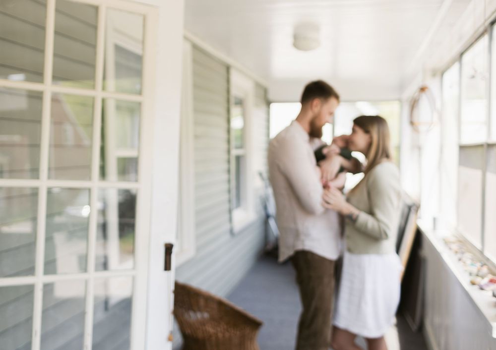 An out of focus shot of a couple holding a newborn on a farmhouse porch with glass windows for a lifestyle newborn photo shoot in Berwick.