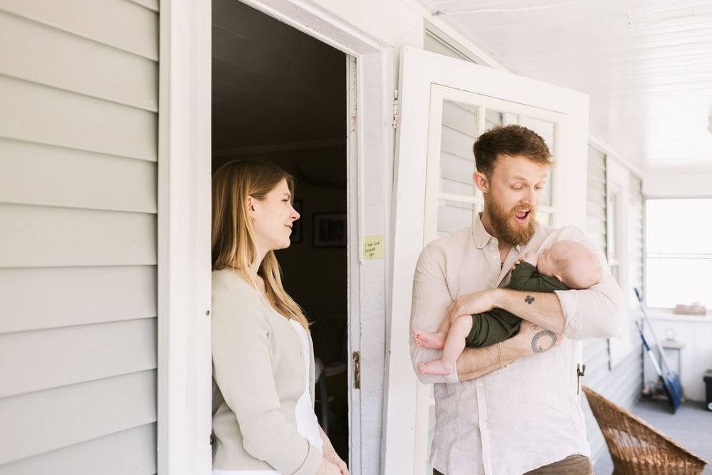 A father holds his newborn son on their porch while the mother looks on during a family photo shoot in Southern Maine. North Shore MA