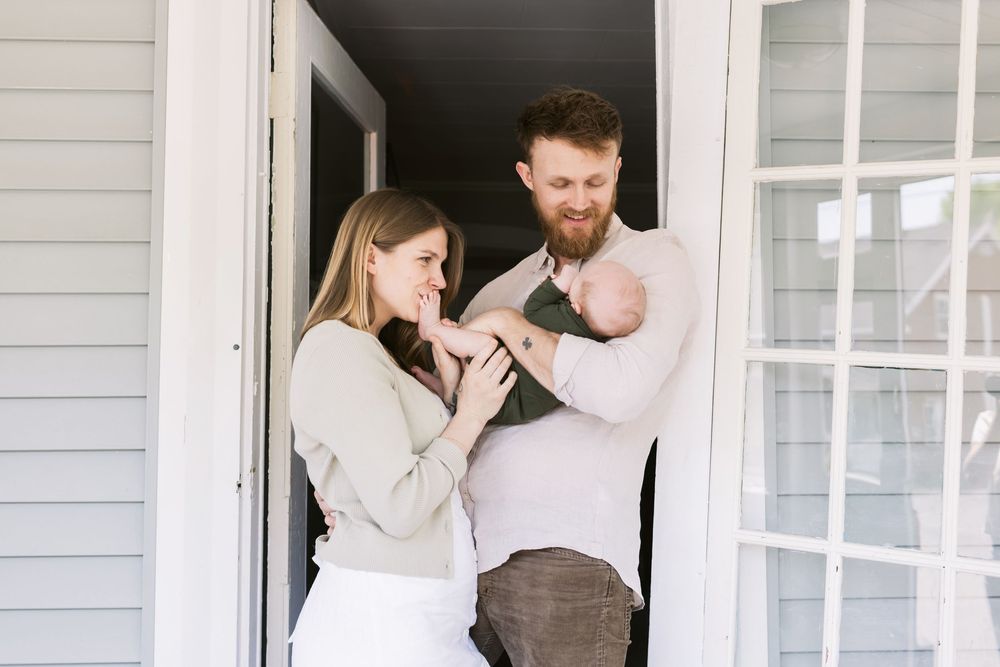 A mother with blond hair kisses her newborn son's feet in the doorway of their porch for a newborn home photo shoot. Lakes Region NH photos
