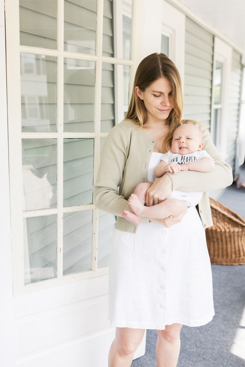 A mother wearing a white dress holds her newborn son on her brightly lit porch during a newborn photo shoot in southern Maine.