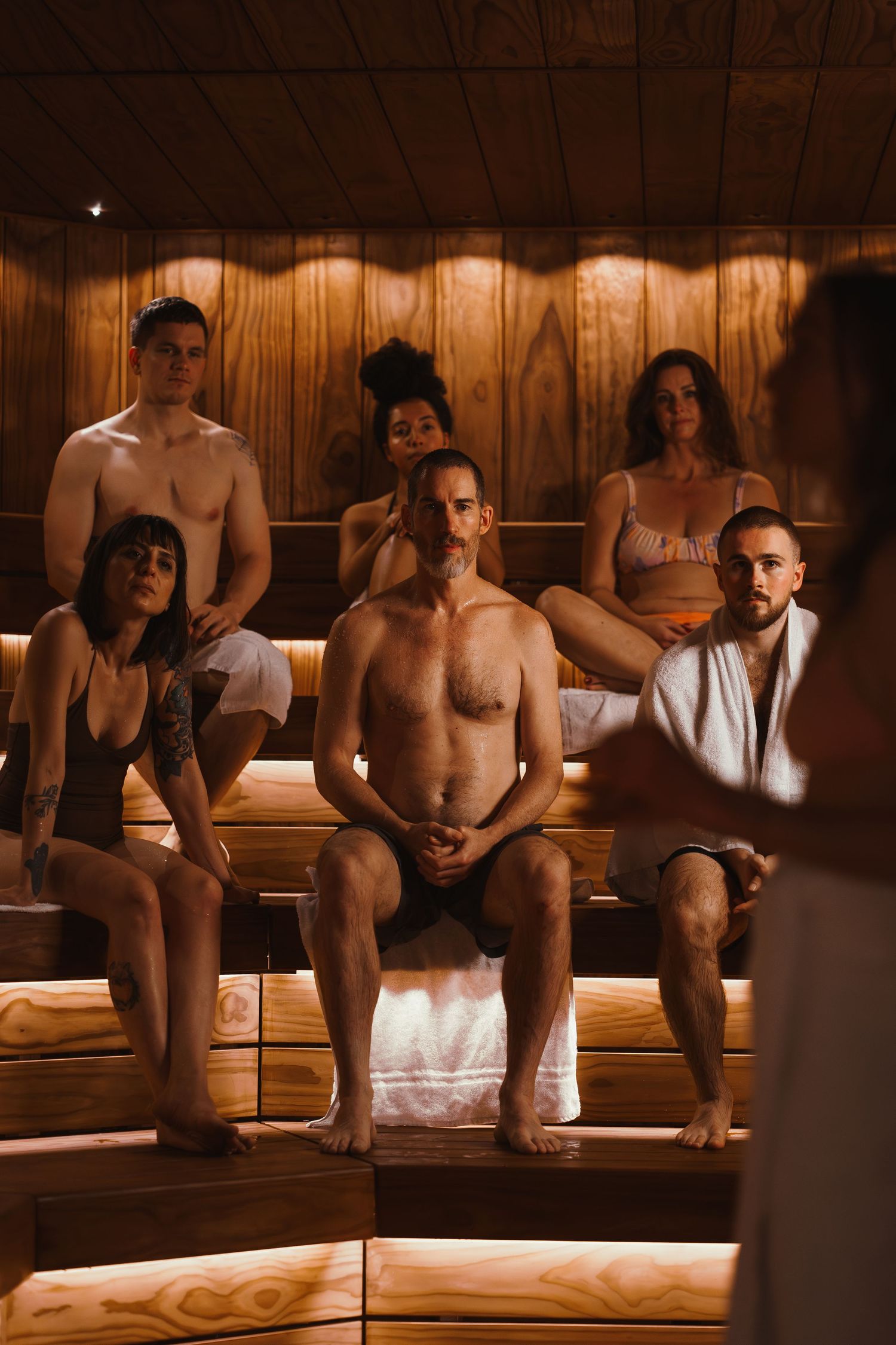 A dimly lit sauna or spa room with wooden walls and benches showing multiple people relaxing in warm tones.