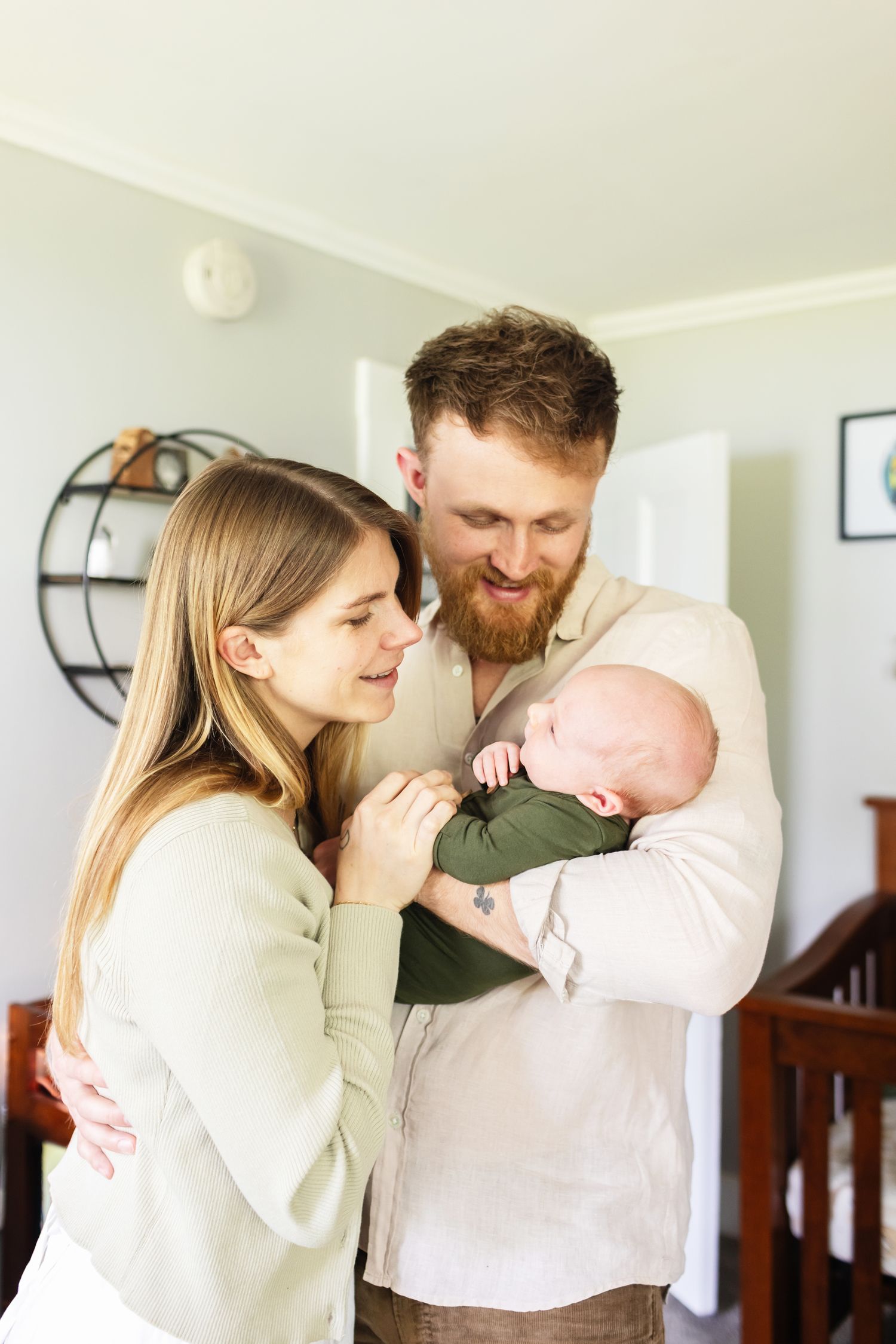 A happy mother and father gazes at their newborn son wearing forest green for newborn photos in the nursery in Berwick, Maine.