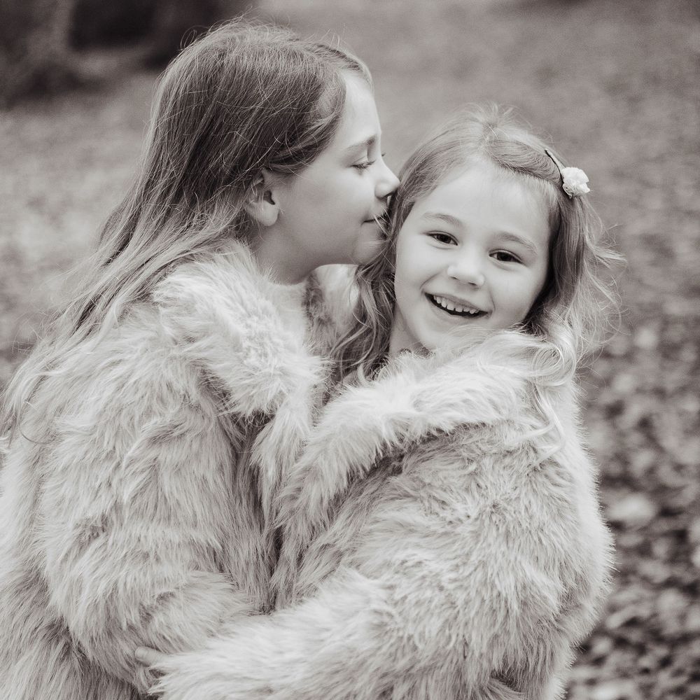 Two young sister hugging. One is laughing to the camera and the other is kissing her younger sisters head.