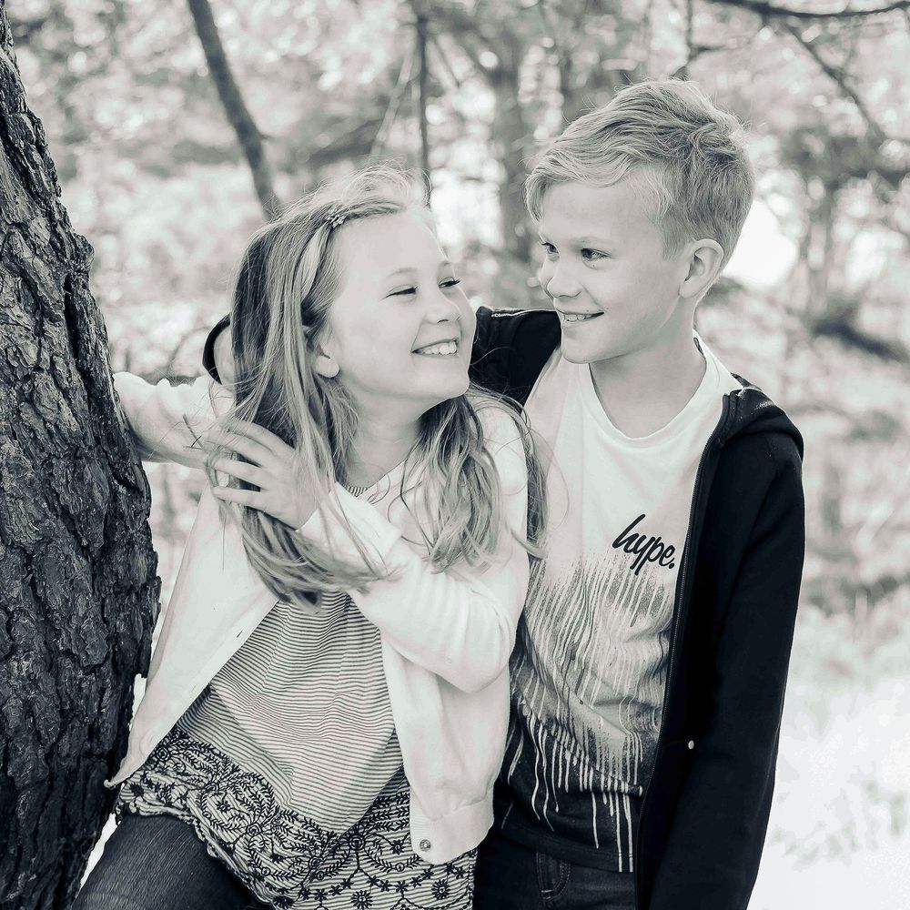 Black and white image of a brother and sister looking at each other laughing by a tree.