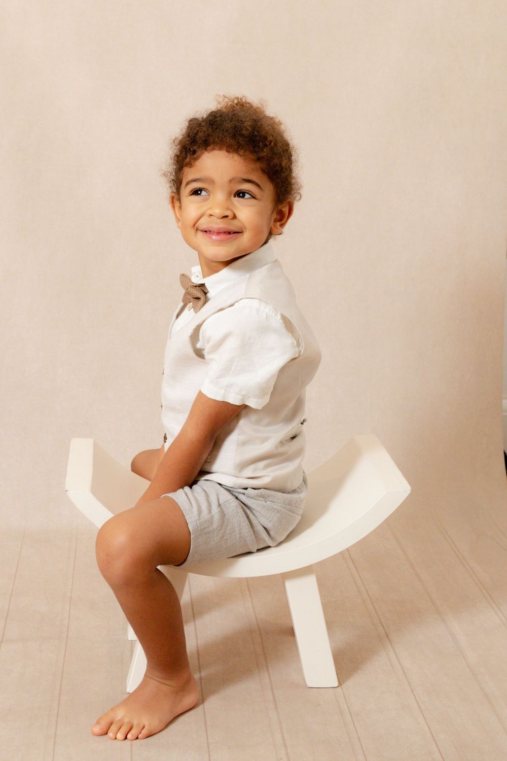 Young boy in a cream studio setting wearing tailored shorts with a waistcoat and bow tie.  Sitting on a white curved chair smiling.